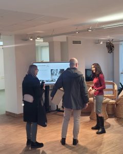 Three people engage in a discussion in a modern museum setting, with a digital display showcasing information about the MATOMI project. The individuals include a woman in a black coat and grey pants, a man in a grey jacket holding a box, and a woman in a red top and skirt, highlighting the project's focus on enhancing visitor experiences.
