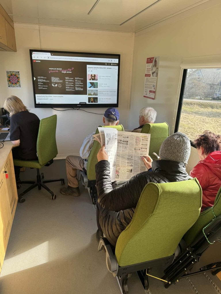 Group of individuals engaging with digital content on a large screen inside a mobile unit, highlighting the inclusive nature of the project. One person reads a newspaper while others observe the display, showcasing community collaboration and continued learning. Group of individuals engaging with digital content on a large screen inside a mobile unit, highlighting the inclusive nature of the project. One person reads a newspaper while others observe the display, showcasing community collaboration and continued learning.