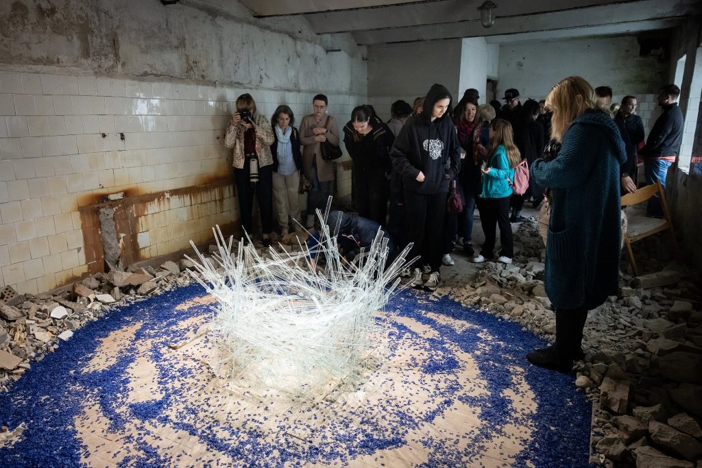 Visitors observe an art installation made of glass and blue materials at the JEWEL Art Residency in Žagarė, showcasing creativity in a rustic, partially ruined space. Visitors observe an art installation made of glass and blue materials at the JEWEL Art Residency in Žagarė, showcasing creativity in a rustic, partially ruined space.