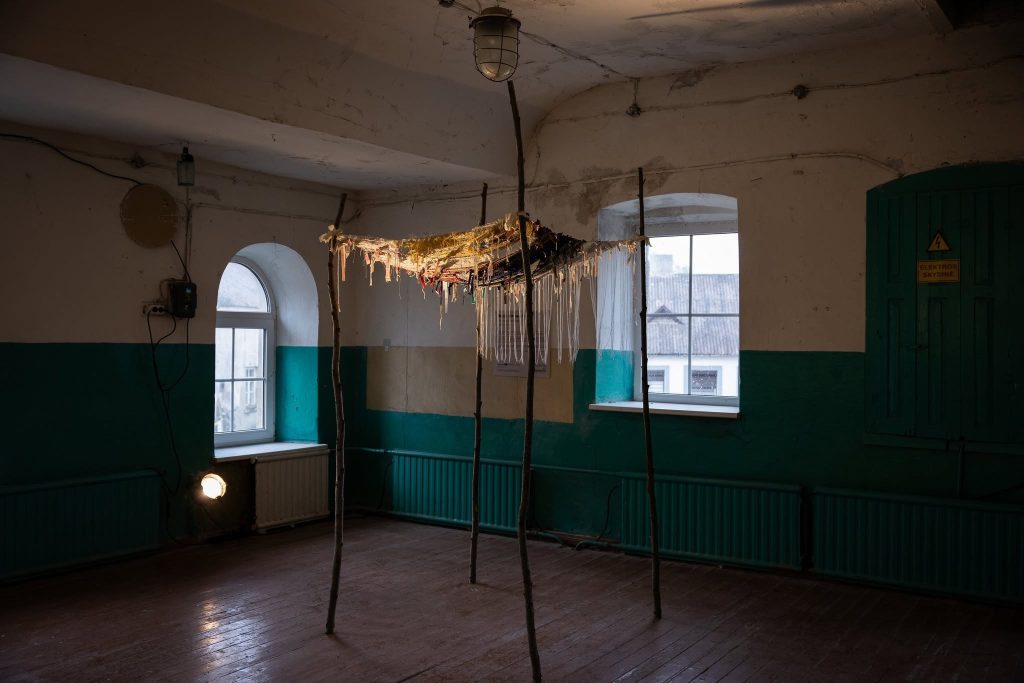 Art installation suspended in an empty room with teal and white walls, featuring a structure made of sticks and various materials, showcasing the creative environment of the JEWEL Art Residency in Žagarė. Natural light streams through the windows, illuminating the unique artwork. Art installation suspended in an empty room with teal and white walls, featuring a structure made of sticks and various materials, showcasing the creative environment of the JEWEL Art Residency in Žagarė. Natural light streams through the windows, illuminating the unique artwork.