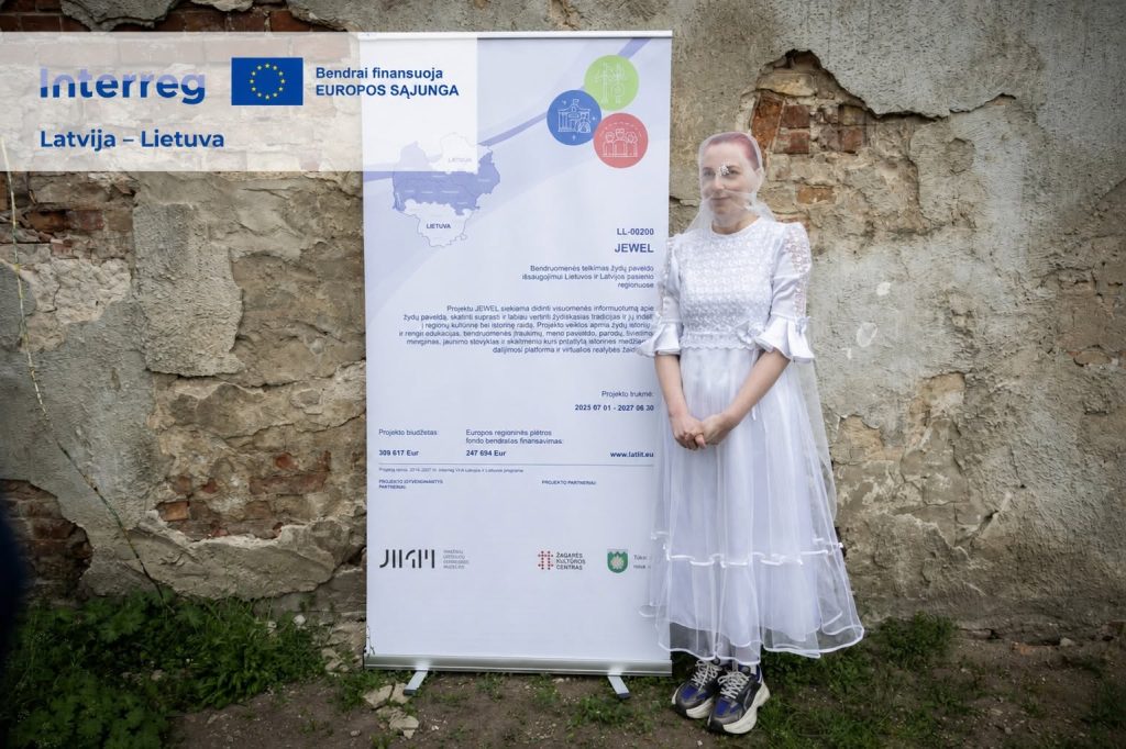 A participant in the JEWEL Art Residency stands beside an informational banner in Žagarė, showcasing the event's focus on cultural collaboration between Latvia and Lithuania. The backdrop features a weathered wall, enhancing the artistic atmosphere of the residency. A participant in the JEWEL Art Residency stands beside an informational banner in Žagarė, showcasing the event's focus on cultural collaboration between Latvia and Lithuania. The backdrop features a weathered wall, enhancing the artistic atmosphere of the residency.