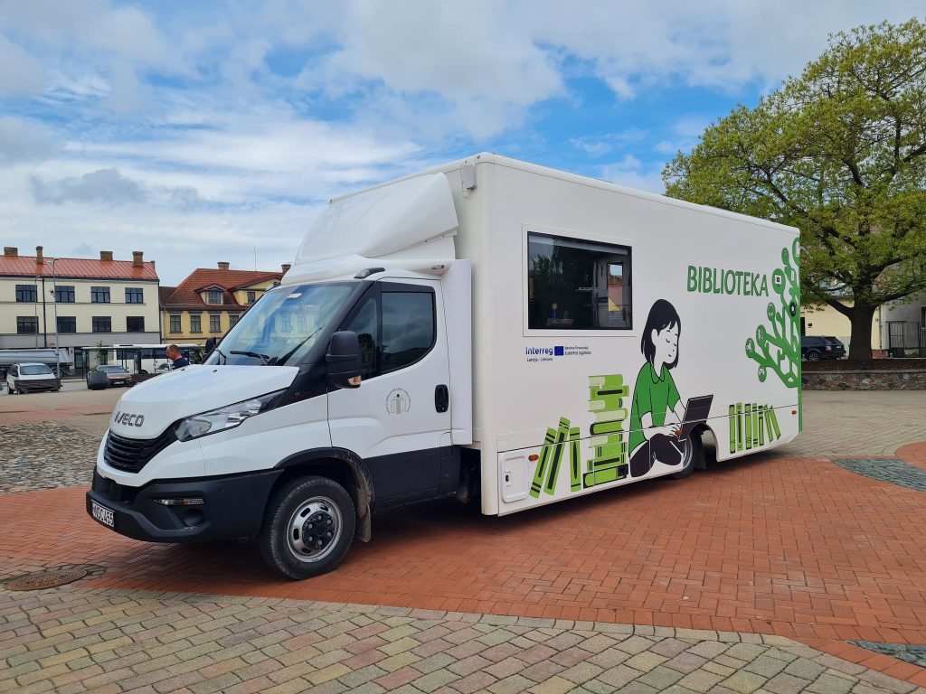 Mobile library vehicle featuring a white exterior with green illustrations, parked on a cobblestone square. The design includes the word "BIBLIOTEKA" and an image of a girl using a laptop, symbolizing community engagement and access to resources. Mobile library vehicle featuring a white exterior with green illustrations, parked on a cobblestone square. The design includes the word "BIBLIOTEKA" and an image of a girl using a laptop, symbolizing community engagement and access to resources.