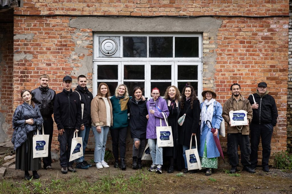 Group photo of JEWEL Art Residency participants in Žagarė, showcasing diverse artists standing in front of a brick building, each holding tote bags, highlighting collaboration and creativity in the art community. Group photo of JEWEL Art Residency participants in Žagarė, showcasing diverse artists standing in front of a brick building, each holding tote bags, highlighting collaboration and creativity in the art community.