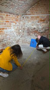 Two individuals measuring a space in a brick-walled room as part of the MATOMI project aimed at enhancing museum visitor experiences.