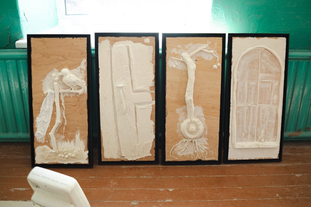 Four textured art panels displayed in a sunlit room, showcasing white sculptural elements on wood backgrounds, part of the JEWEL Art Residency in Žagarė. Four textured art panels displayed in a sunlit room, showcasing white sculptural elements on wood backgrounds, part of the JEWEL Art Residency in Žagarė.