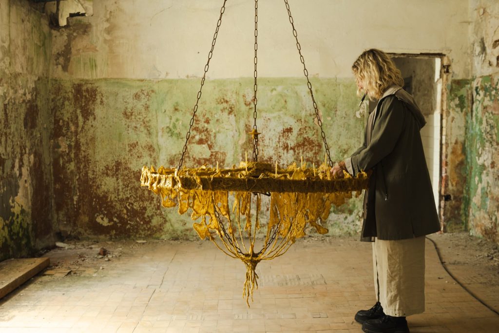 Artist interacting with a unique chandelier made of textured yellow material, set against a backdrop of peeling green walls in an abandoned space, during the JEWEL Art Residency in Žagarė. Artist interacting with a unique chandelier made of textured yellow material, set against a backdrop of peeling green walls in an abandoned space, during the JEWEL Art Residency in Žagarė.