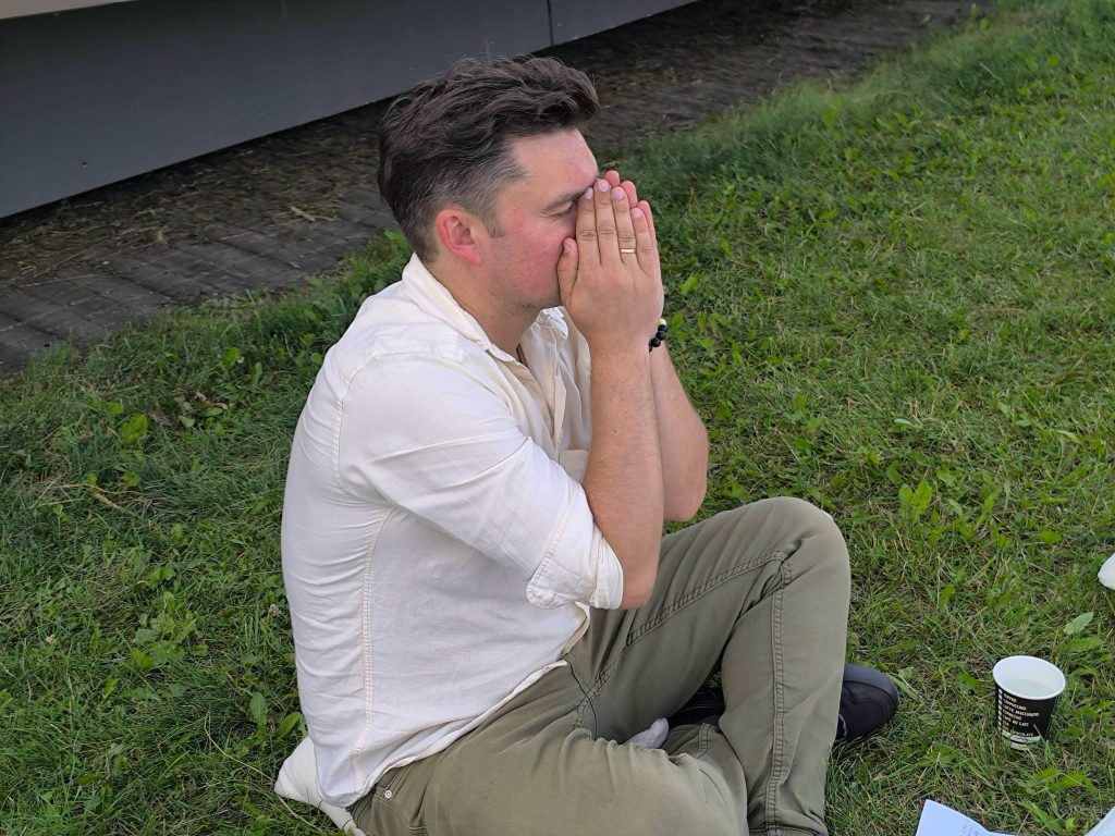 A participant at the "Bee Guide" educational workshop sits on the grass, with hands covering his face, reflecting on the session. Nearby, a paper cup and materials related to the workshop are visible. A participant at the "Bee Guide" educational workshop sits on the grass, with hands covering his face, reflecting on the session. Nearby, a paper cup and materials related to the workshop are visible.
