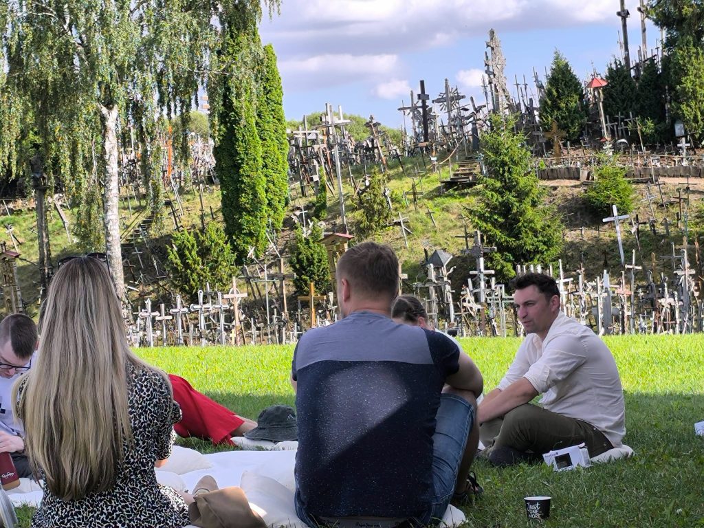 Participants engaged in an educational workshop titled "Bee Guide," seated on the grass with a backdrop of a hillside covered in numerous crosses and trees, illustrating a unique blend of nature and history. Participants engaged in an educational workshop titled "Bee Guide," seated on the grass with a backdrop of a hillside covered in numerous crosses and trees, illustrating a unique blend of nature and history.