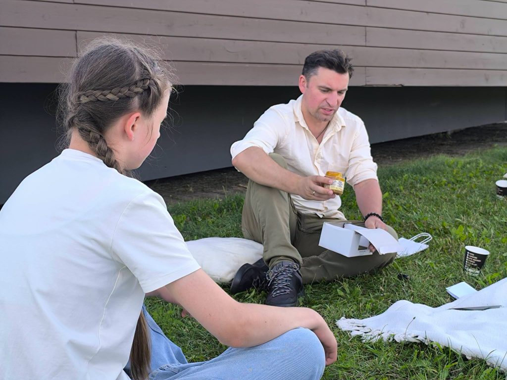 Participants engage in an educational workshop titled "Bee Guide," with a focus on beekeeping techniques. A man demonstrates the use of bee-related products to a young girl, as they sit on the grass surrounded by materials related to the workshop. Participants engage in an educational workshop titled "Bee Guide," with a focus on beekeeping techniques. A man demonstrates the use of bee-related products to a young girl, as they sit on the grass surrounded by materials related to the workshop.