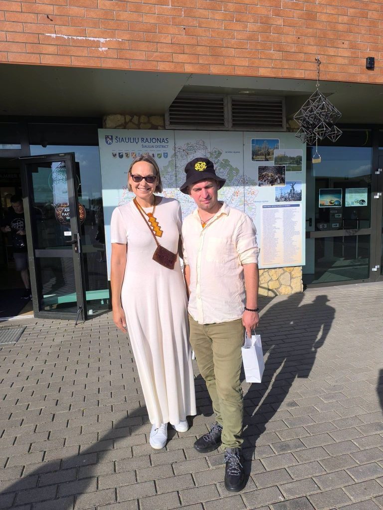 Two participants of the "Bee Guide" educational workshop stand outside a building in Šiauliai District, smiling for the camera. The woman is wearing a long white dress and sunglasses, while the man is in a light shirt and green pants, holding a small shopping bag. A map of the district is visible in the background, highlighting local attractions. Two participants of the "Bee Guide" educational workshop stand outside a building in Šiauliai District, smiling for the camera. The woman is wearing a long white dress and sunglasses, while the man is in a light shirt and green pants, holding a small shopping bag. A map of the district is visible in the background, highlighting local attractions.
