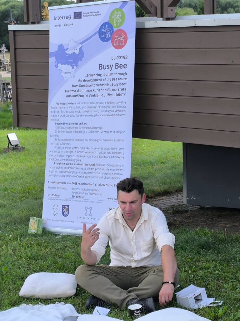 Man sitting on the grass at an educational workshop titled "Bee Guide," discussing the development of the Bee route from Kuršēni to Ventspils, with an informational banner in the background detailing the project and its goals for enhancing tourism. Man sitting on the grass at an educational workshop titled "Bee Guide," discussing the development of the Bee route from Kuršēni to Ventspils, with an informational banner in the background detailing the project and its goals for enhancing tourism.