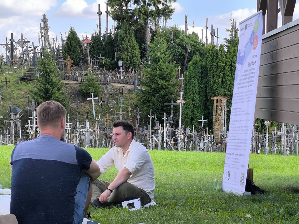Participants engage in an educational workshop titled "Bee Guide," focusing on bee conservation and ecology, set against a backdrop of a unique landscape featuring numerous crosses and trees. Participants engage in an educational workshop titled "Bee Guide," focusing on bee conservation and ecology, set against a backdrop of a unique landscape featuring numerous crosses and trees.