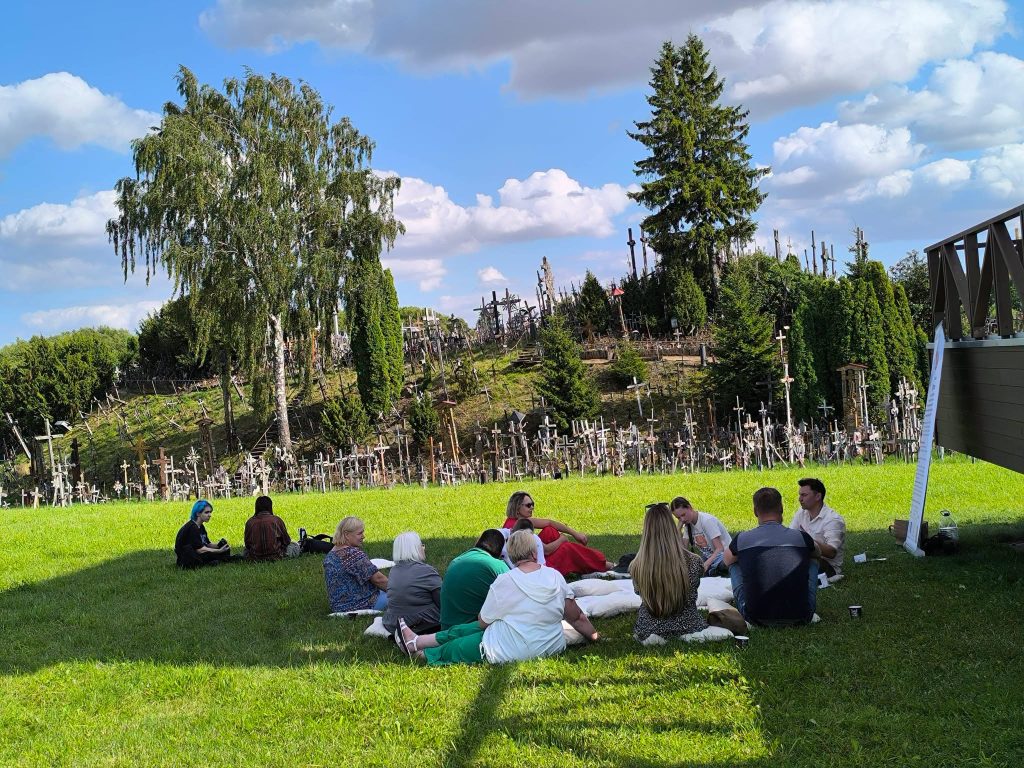 Participants engaged in an educational workshop titled "Bee Guide" sit on the grass, surrounded by a scenic landscape featuring a hill adorned with numerous crosses and trees under a blue sky with fluffy clouds. Participants engaged in an educational workshop titled "Bee Guide" sit on the grass, surrounded by a scenic landscape featuring a hill adorned with numerous crosses and trees under a blue sky with fluffy clouds.