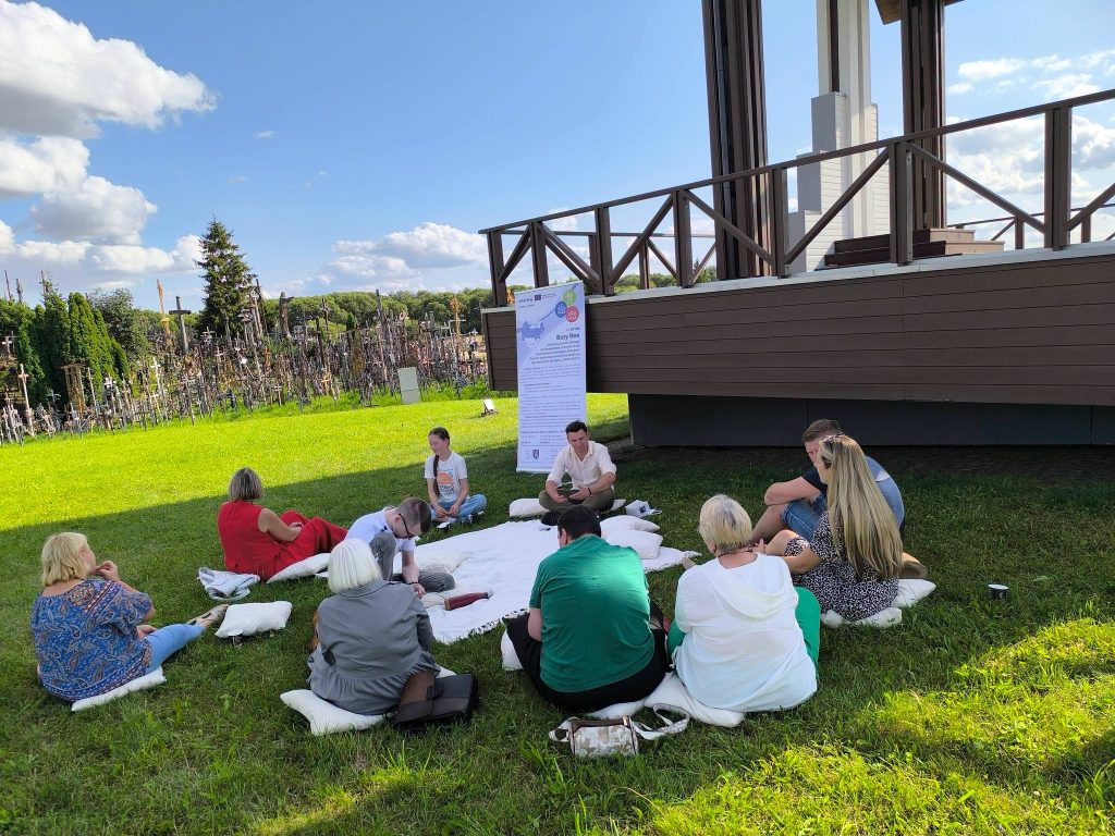 Participants engaging in the "Bee Guide" educational workshop, seated on cushions in a grassy area, with a banner in the background highlighting workshop details and a backdrop of trees and wooden structures. Participants engaging in the "Bee Guide" educational workshop, seated on cushions in a grassy area, with a banner in the background highlighting workshop details and a backdrop of trees and wooden structures.