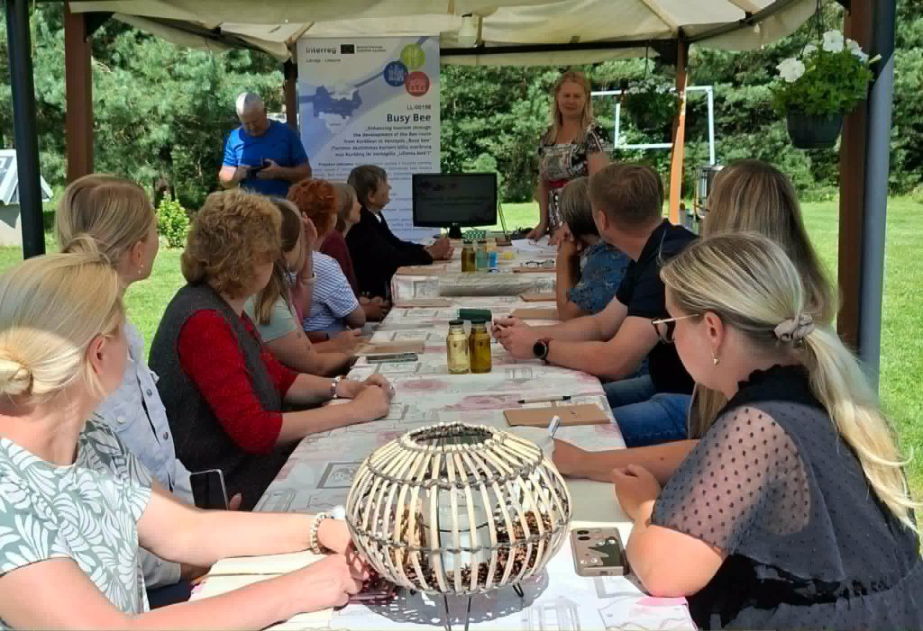 Participants engaged in an educational workshop on creating solid perfumes using scented wax, seated at a table outdoors under a canopy. A presenter is demonstrating techniques while attendees take notes and observe. The setting includes a display banner and various materials for the workshop.