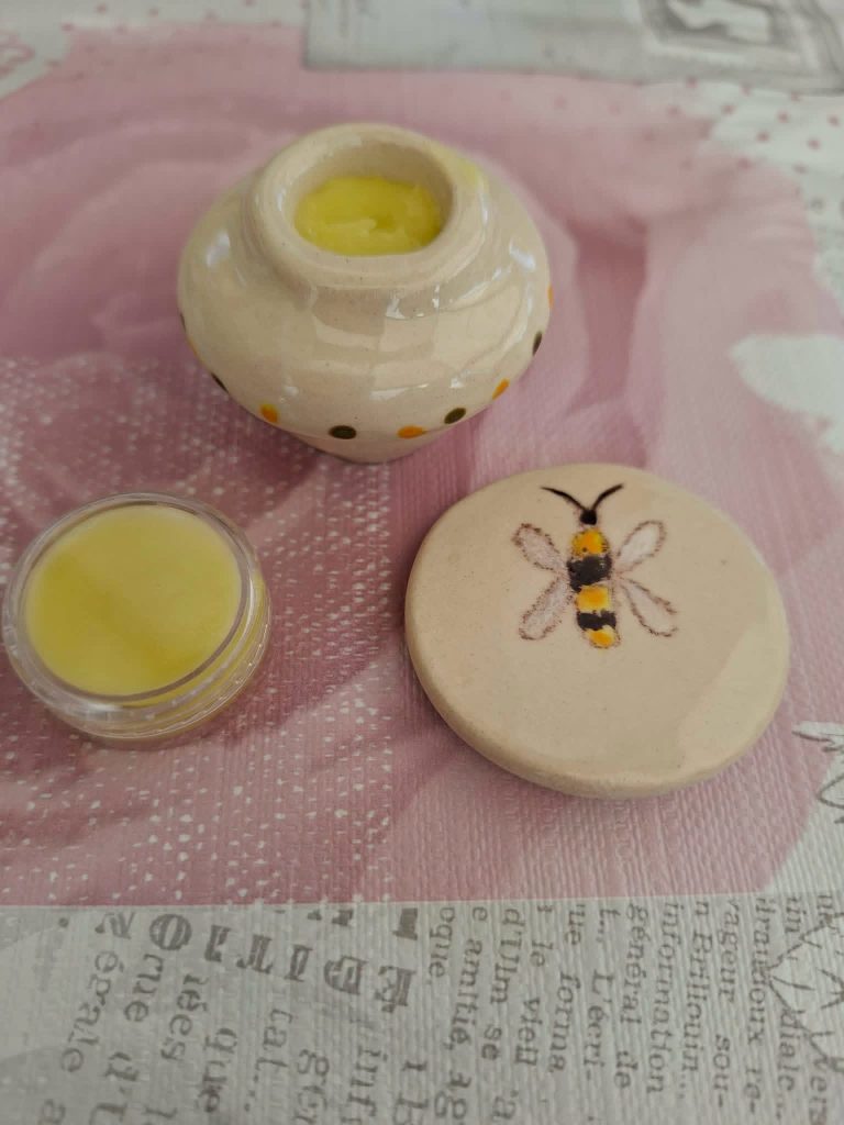 Close-up of a handmade ceramic container filled with solid perfume, alongside a small lip balm and a decorative lid featuring a bee illustration, set against a textured background.