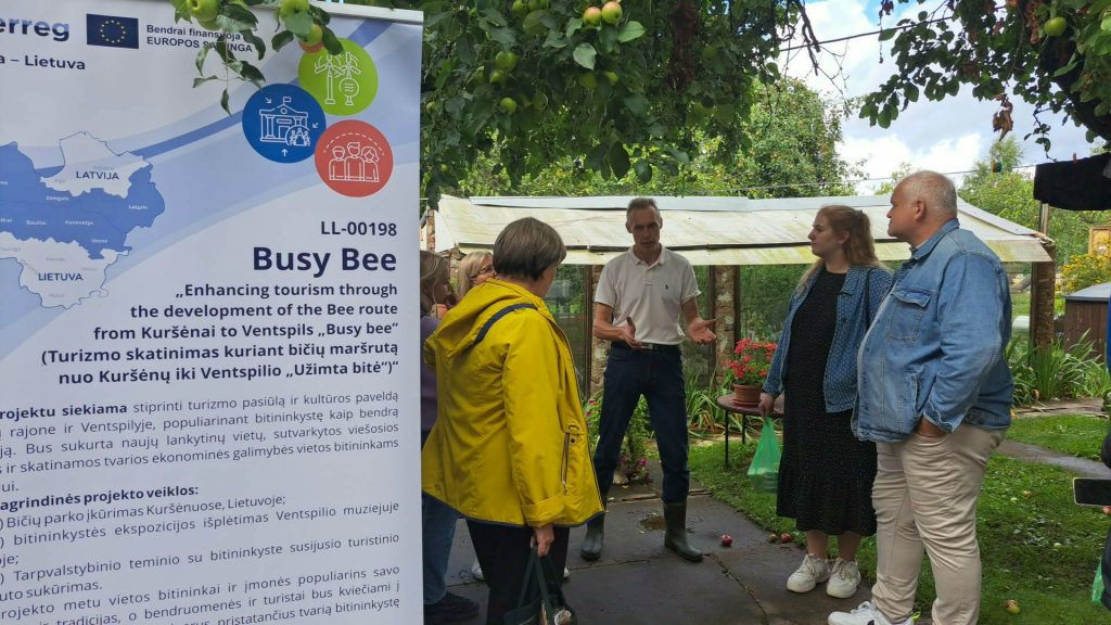 Participants engaging in an educational workshop titled "Bee Therapy," discussing the development of a bee tourism route from Kuršėnai to Ventspils, with a focus on local cultural heritage and economic opportunities. The setting features a banner outlining the project and a backdrop of greenery.