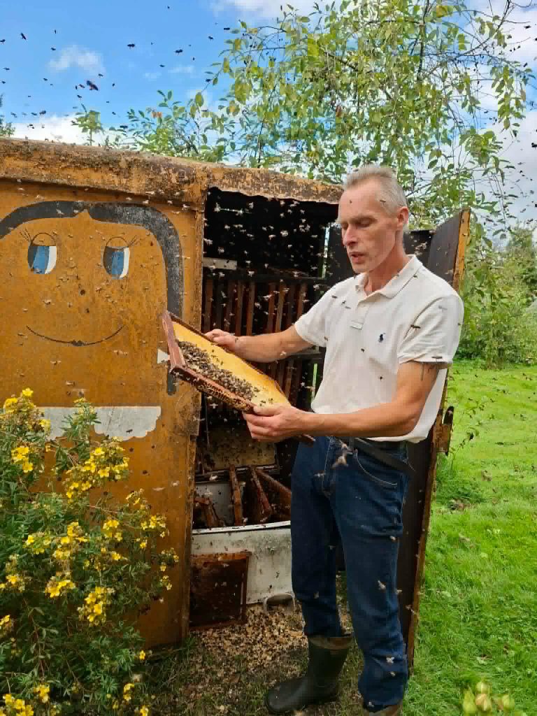 A beekeeper holding a honeycomb frame filled with bees, standing in front of a colorful beehive with a smiling face design, surrounded by greenery and flowers during an educational workshop on "Bee Therapy."