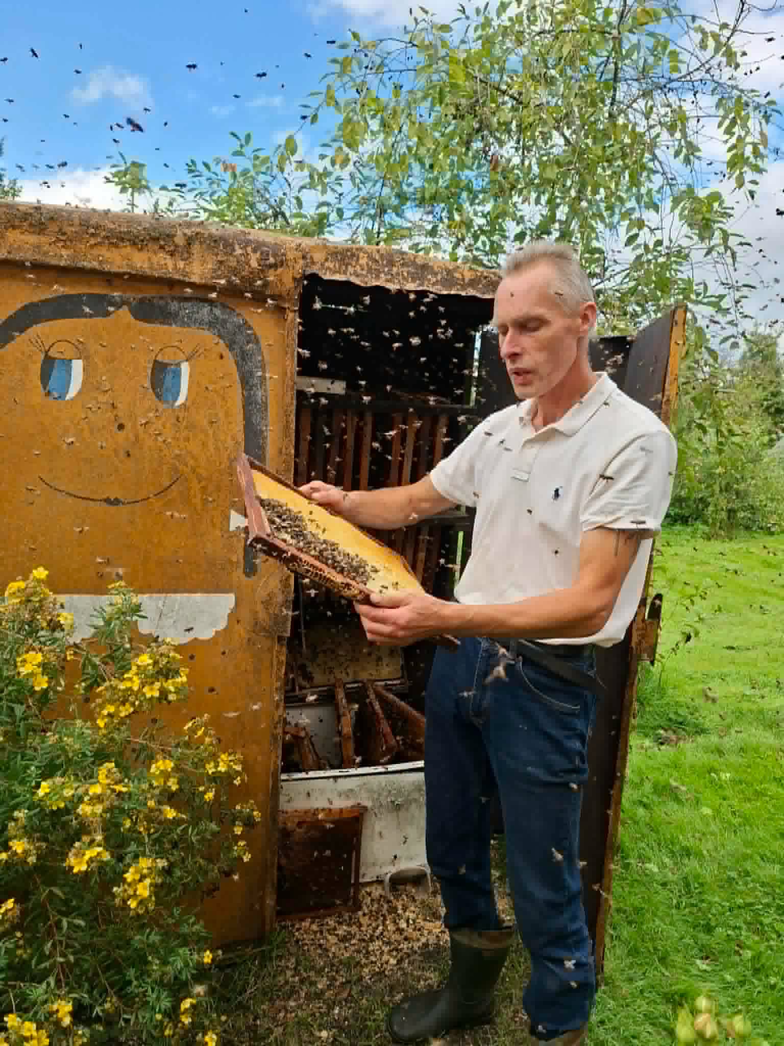 A beekeeper holding a honeycomb frame filled with bees, standing in front of a colorful beehive with a smiling face design, surrounded by greenery and flowers during an educational workshop on "Bee Therapy."