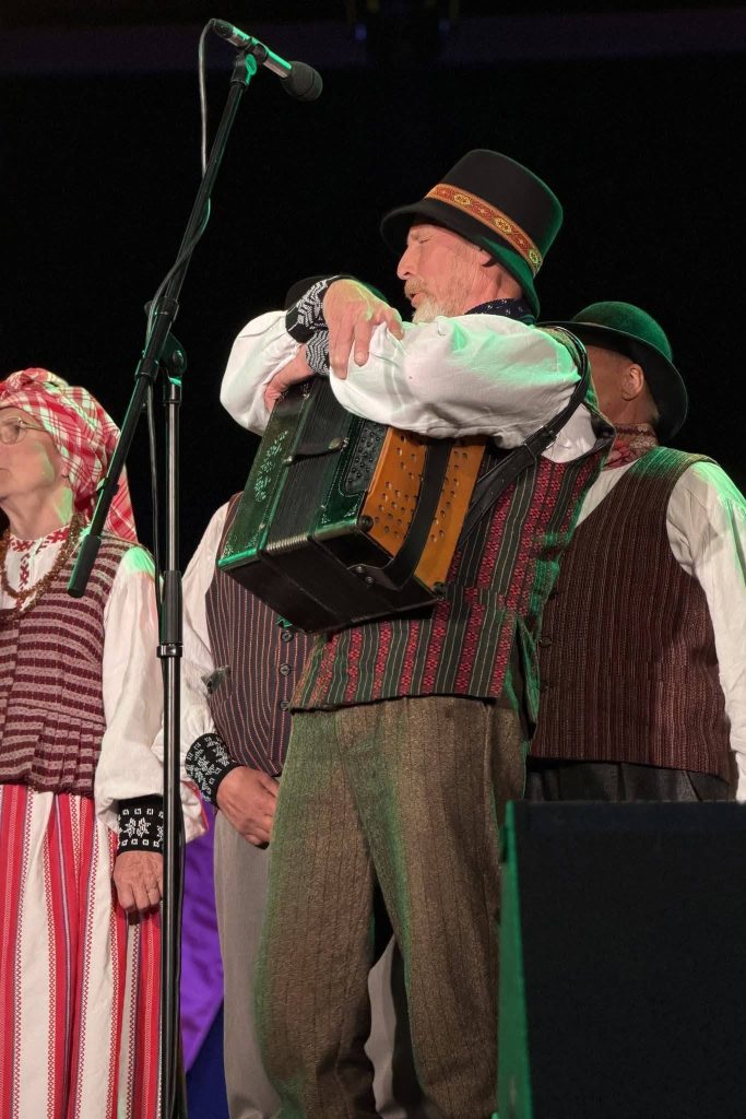 A musician in traditional attire plays the accordion on stage during a cultural performance in Baisogala, showcasing the musical traditions of Lithuania and Latvia, with other performers in the background.