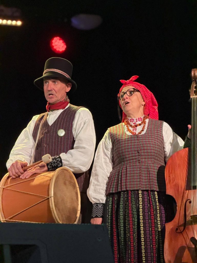 Traditional Lithuanian and Latvian musicians performing on stage in Baisogala, showcasing cultural attire and instruments, including a drum and a stringed instrument, during a vibrant musical event.
