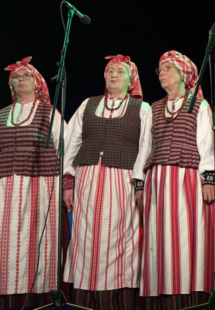 Three women dressed in traditional Latvian folk costumes, featuring red and white striped skirts and vests, perform together on stage during a musical event in Baisogala, celebrating the cultural traditions of Lithuania and Latvia.