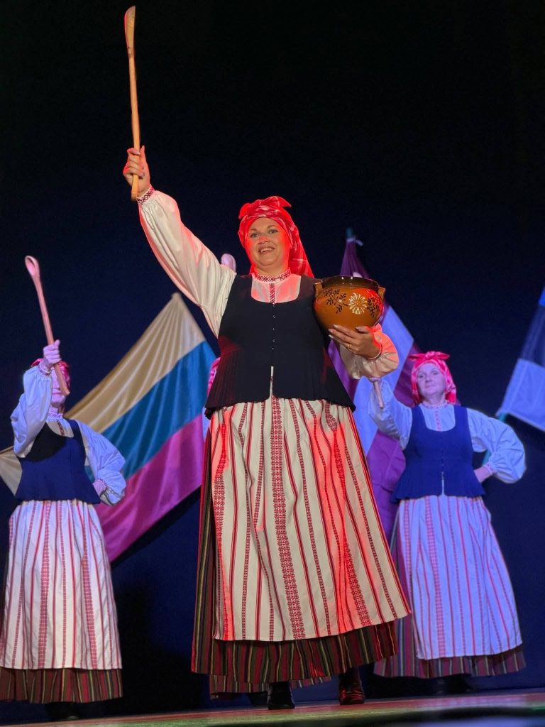 A performer in traditional Lithuanian attire holds a wooden stick and a decorative bowl while leading a cultural dance, with fellow dancers in the background, showcasing the vibrant heritage of Lithuania and Latvia during a musical event in Baisogala.