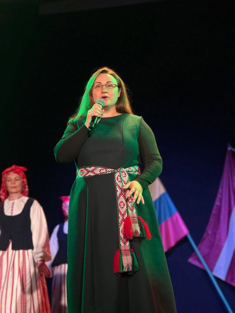 Woman in traditional attire performing on stage during a cultural event, with colorful flags in the background, highlighting the musical collaboration between Lithuanian and Latvian traditions in Baisogala.