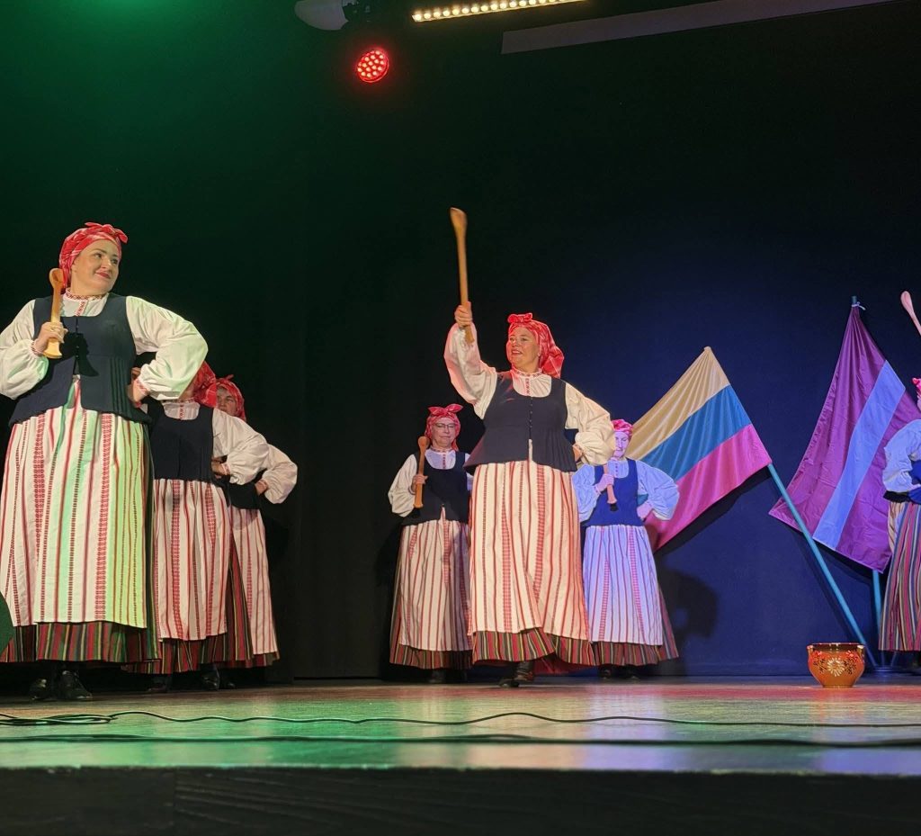 Traditional dancers in colorful costumes perform on stage during a cultural event in Baisogala, showcasing Lithuanian and Latvian traditions. The dancers hold wooden sticks and are surrounded by flags representing both nations, highlighting the fusion of their musical heritage.