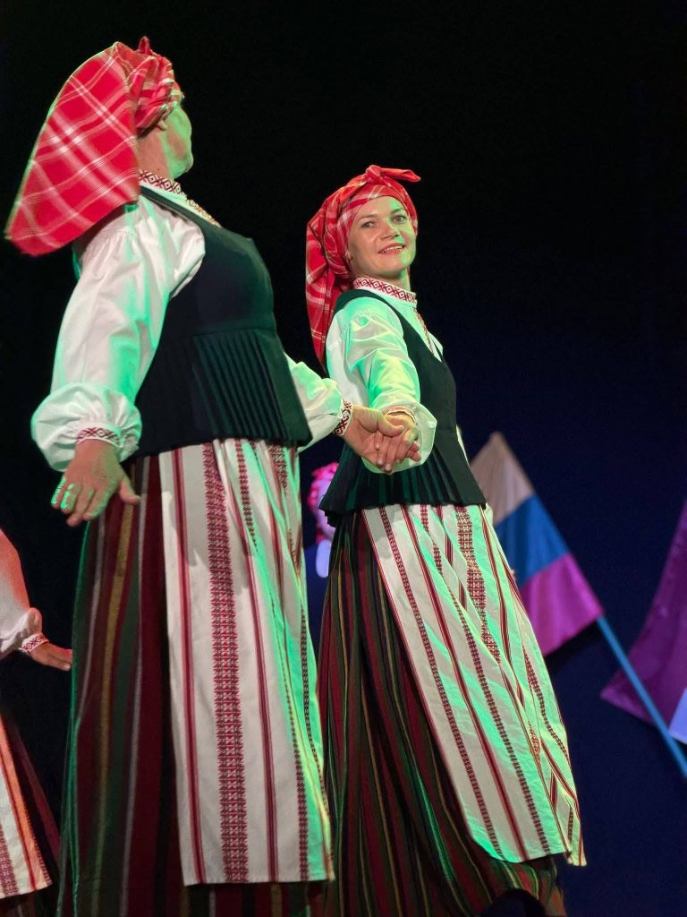Two women in traditional Latvian attire perform a folk dance, showcasing vibrant costumes with intricate patterns. The scene is set against a backdrop of colorful flags, highlighting the cultural connection between Lithuanian and Latvian traditions during a musical event in Baisogala.