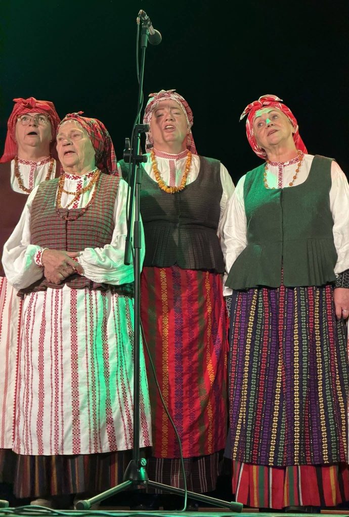 Four women in traditional Latvian attire sing together at a microphone, showcasing their cultural heritage during a musical event in Baisogala, where Lithuanian and Latvian traditions converge.