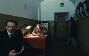 Two men engaged in conversation in a dimly lit room, one in a suit and the other in a military uniform, seated at a table with a red tablecloth. The setting features a vintage lamp, a typewriter, and a glass of water, reflecting a historical context related to the MATOMI project aimed at enhancing museum visitor experiences.