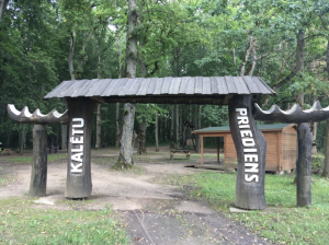 Entrance to Kalēti Forest Park featuring wooden archway with carved signage, surrounded by lush greenery, inviting visitors to explore biodiversity during the educational walk. Entrance to Kalēti Forest Park featuring wooden archway with carved signage, surrounded by lush greenery, inviting visitors to explore biodiversity during the educational walk.