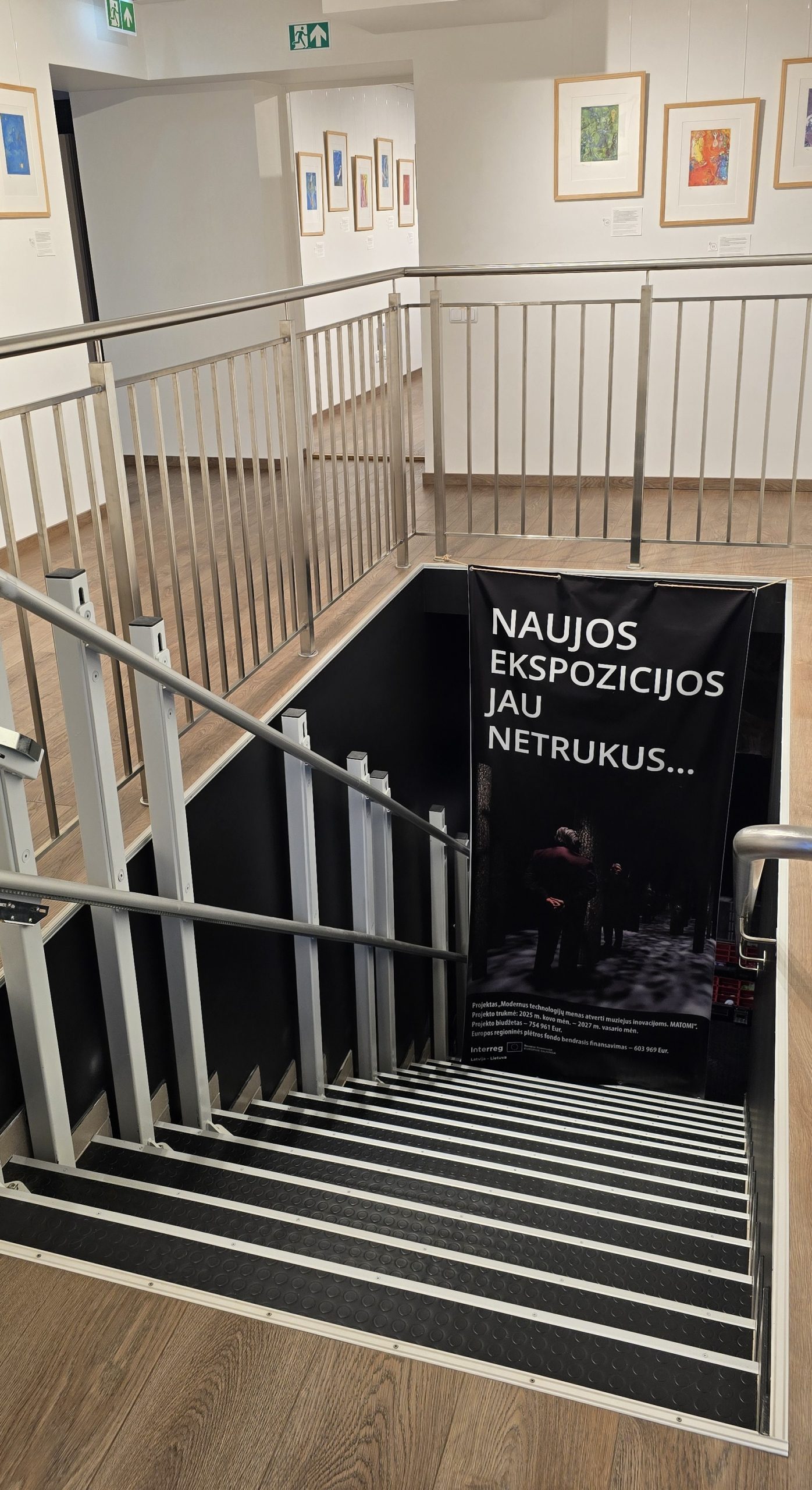 Staircase leading to a new exhibition area in a museum, featuring a banner announcing upcoming exhibits. The space is well-lit with framed artworks displayed on the walls, highlighting the transition to modern visitor experiences in the MATOMI project.