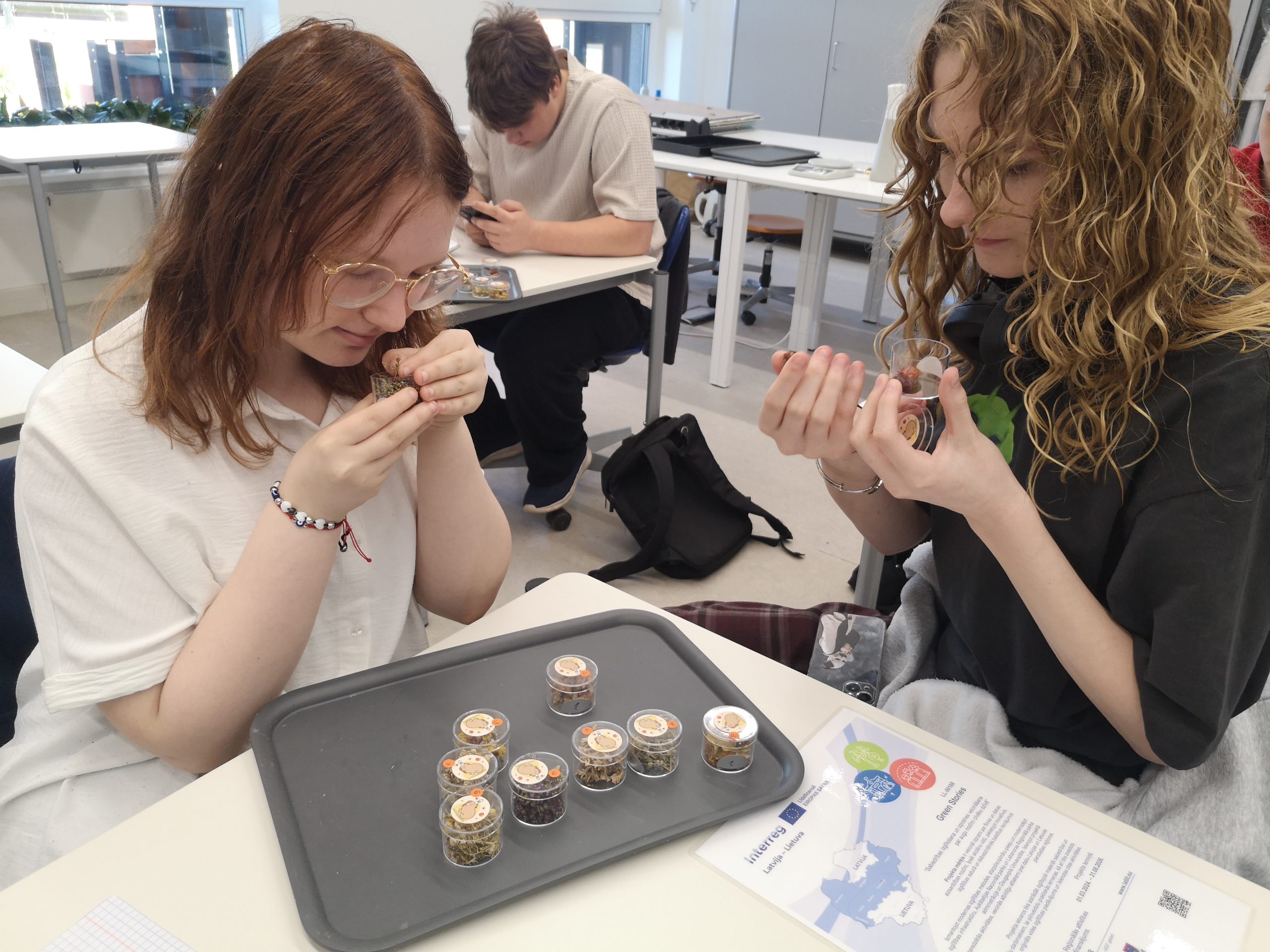Students engaged in a hands-on educational activity, exploring various natural materials in transparent containers during the Green Stories Project. One student examines a sample closely while another observes their own container, highlighting the project's focus on nature exploration and innovative learning experiences. Students engaged in a hands-on educational activity, exploring various natural materials in transparent containers during the Green Stories Project. One student examines a sample closely while another observes their own container, highlighting the project's focus on nature exploration and innovative learning experiences.