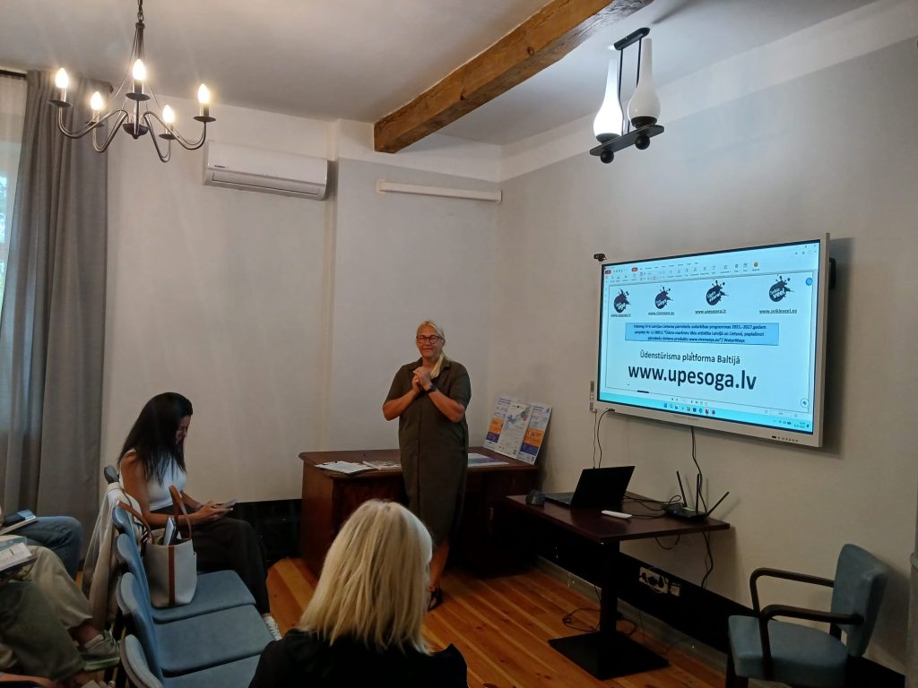 Image of a seminar on water tourism trends in Latgale, held at Lielborne Manor. A speaker presents in front of a screen displaying information about the event, while attendees engage in discussion. The room features a chandelier and wooden beams, creating a cozy atmosphere.