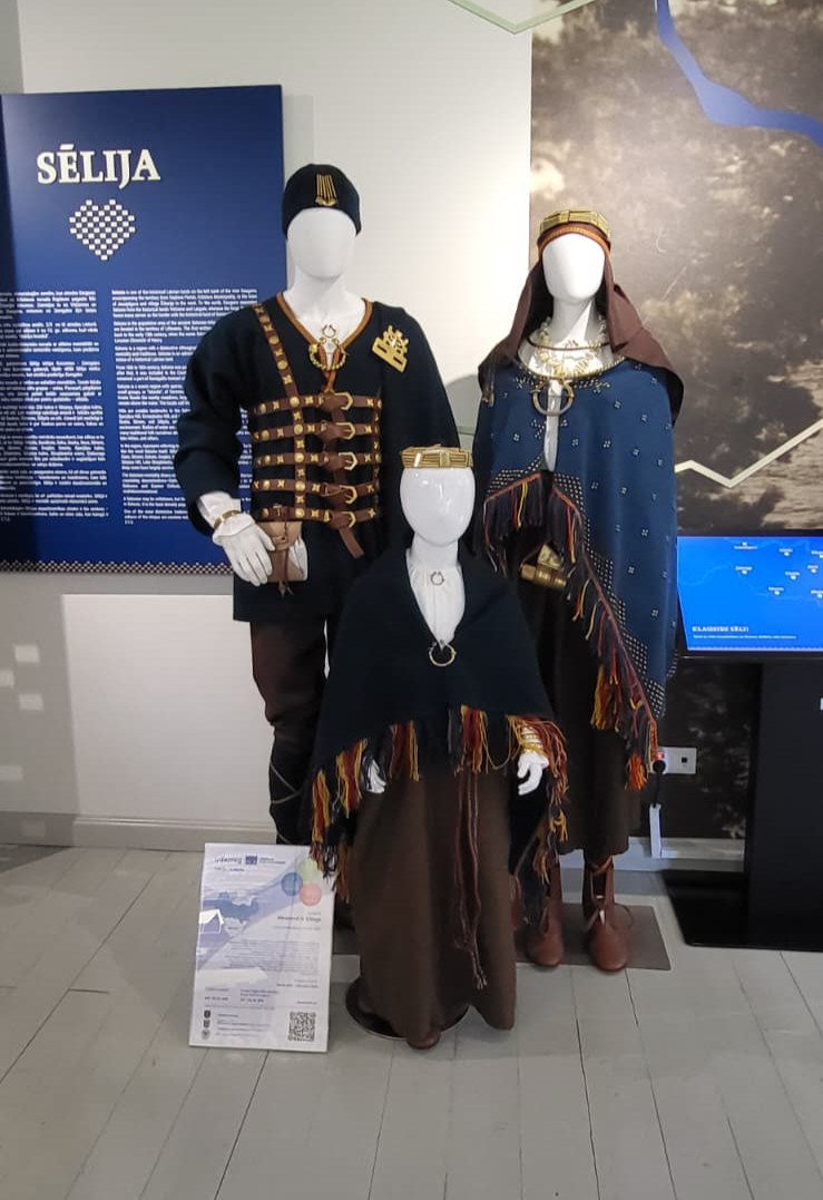 Display of traditional Sēlija folk costumes and archaeological jewelry, featuring three mannequins dressed in intricately designed outfits at the "Sēļu sēta" exhibition, highlighting the revival of cultural heritage.