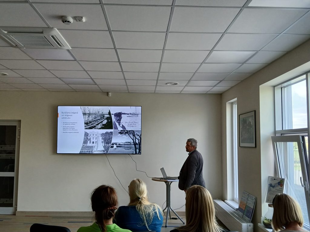 A speaker presents at the "Water Tourism Trends in Zemgale" seminar in Jelgava, with a large screen displaying informative slides about water tourism, while attendees listen attentively. A speaker presents at the "Water Tourism Trends in Zemgale" seminar in Jelgava, with a large screen displaying informative slides about water tourism, while attendees listen attentively.