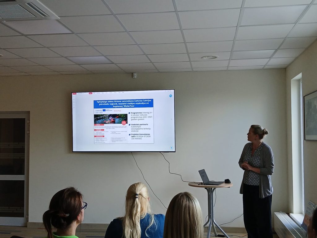 Speaker presenting at the "Water Tourism Trends in Zemgale" seminar in Jelgava, with a projector displaying information about the Interreg program and tourism development initiatives. Attendees are engaged in the presentation. Speaker presenting at the "Water Tourism Trends in Zemgale" seminar in Jelgava, with a projector displaying information about the Interreg program and tourism development initiatives. Attendees are engaged in the presentation.