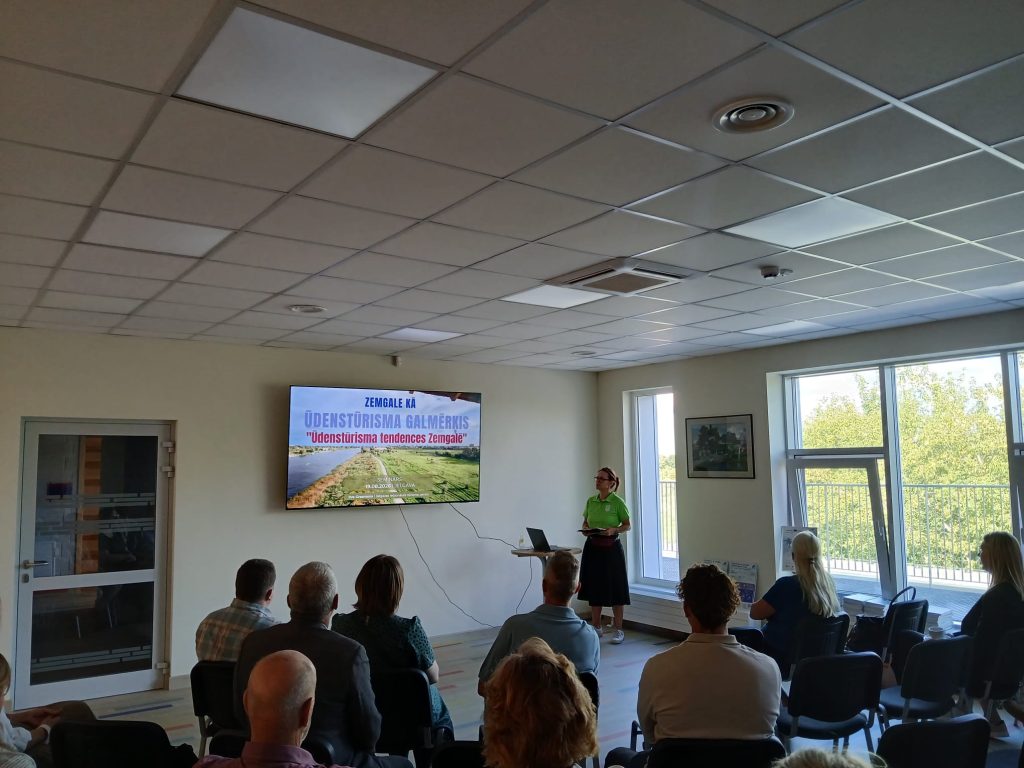 Seminar on water tourism trends in Zemgale, featuring a speaker in a green shirt presenting in front of an audience, with a large screen displaying the seminar title. The setting is a well-lit room with large windows and a professional atmosphere. Seminar on water tourism trends in Zemgale, featuring a speaker in a green shirt presenting in front of an audience, with a large screen displaying the seminar title. The setting is a well-lit room with large windows and a professional atmosphere.
