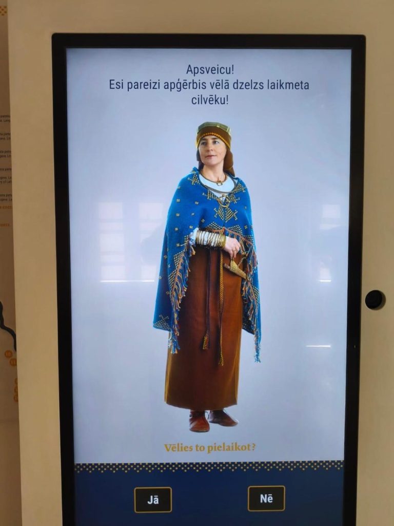Interactive Magic Mirror displaying a woman in traditional medieval attire, featuring a blue cape and brown dress, inviting users to engage with the exhibit. The screen prompts, "Would you like to try it on?" in Latvian.