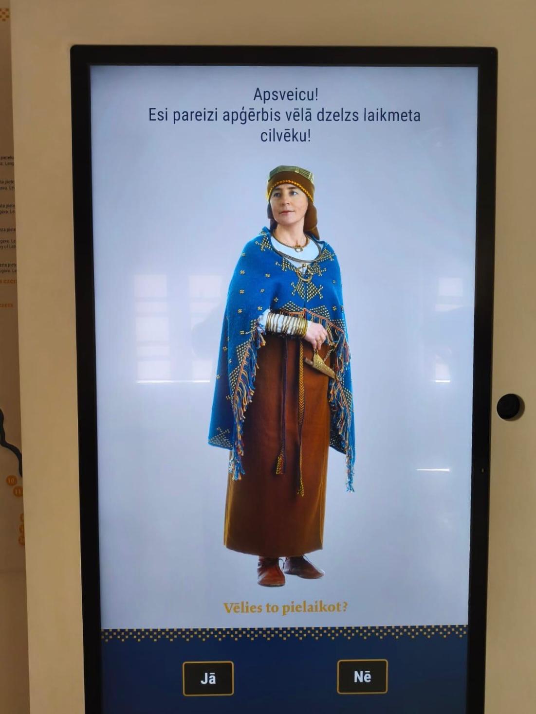 Interactive Magic Mirror displaying a woman in traditional medieval attire, featuring a blue cape and brown dress, inviting users to engage with the exhibit. The screen prompts, "Would you like to try it on?" in Latvian.