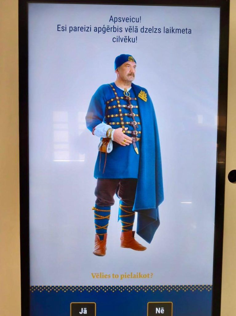 Interactive Magic Mirror displaying a man in historical blue attire with decorative elements, inviting users to try on traditional clothing. The screen features prompts in Latvian language for user interaction.