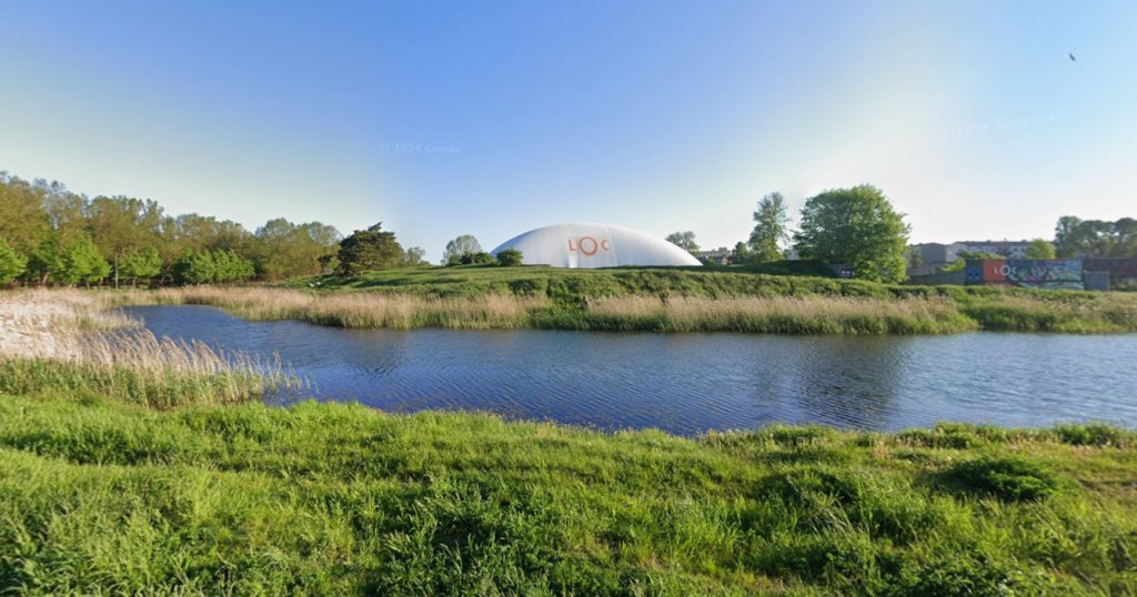 Scenic view of a freshwater ecosystem in Lithuania, featuring a calm water body surrounded by lush greenery and tall grasses, with a distinctive dome structure in the background, highlighting the Rewater project aimed at ecosystem restoration.