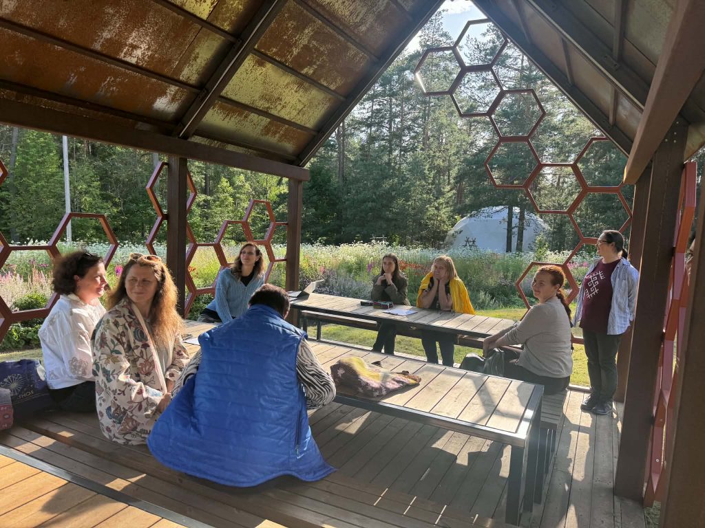 Group of people engaged in discussion under a hexagonal pavilion surrounded by nature, part of the Green Stories Project focused on educational experiences and nature exploration. Group of people engaged in discussion under a hexagonal pavilion surrounded by nature, part of the Green Stories Project focused on educational experiences and nature exploration.