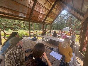 Participants engaged in a collaborative educational session under a unique hexagonal pavilion surrounded by nature, as part of the Green Stories Project. The setting features a rustic design with open seating, promoting interaction and learning about environmental solutions.