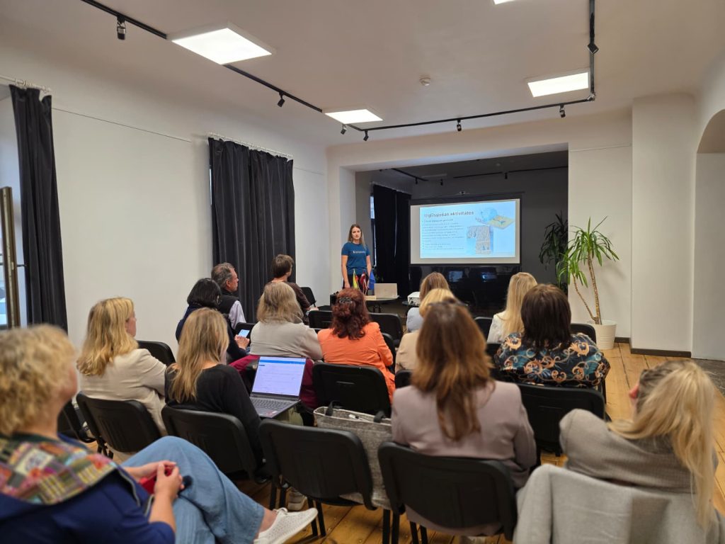 Audience attending a seminar in Saldus focused on the Geosites database, with a presenter discussing practical applications. The room features a screen displaying relevant information, and attendees are engaged and taking notes.