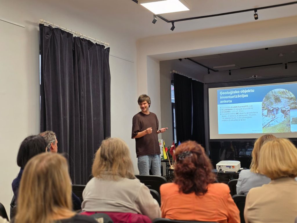A speaker presents at a seminar in Saldus, discussing the Geosites database and its practical applications, with an audience engaged in the session. A presentation slide showcasing geological survey information is visible in the background.