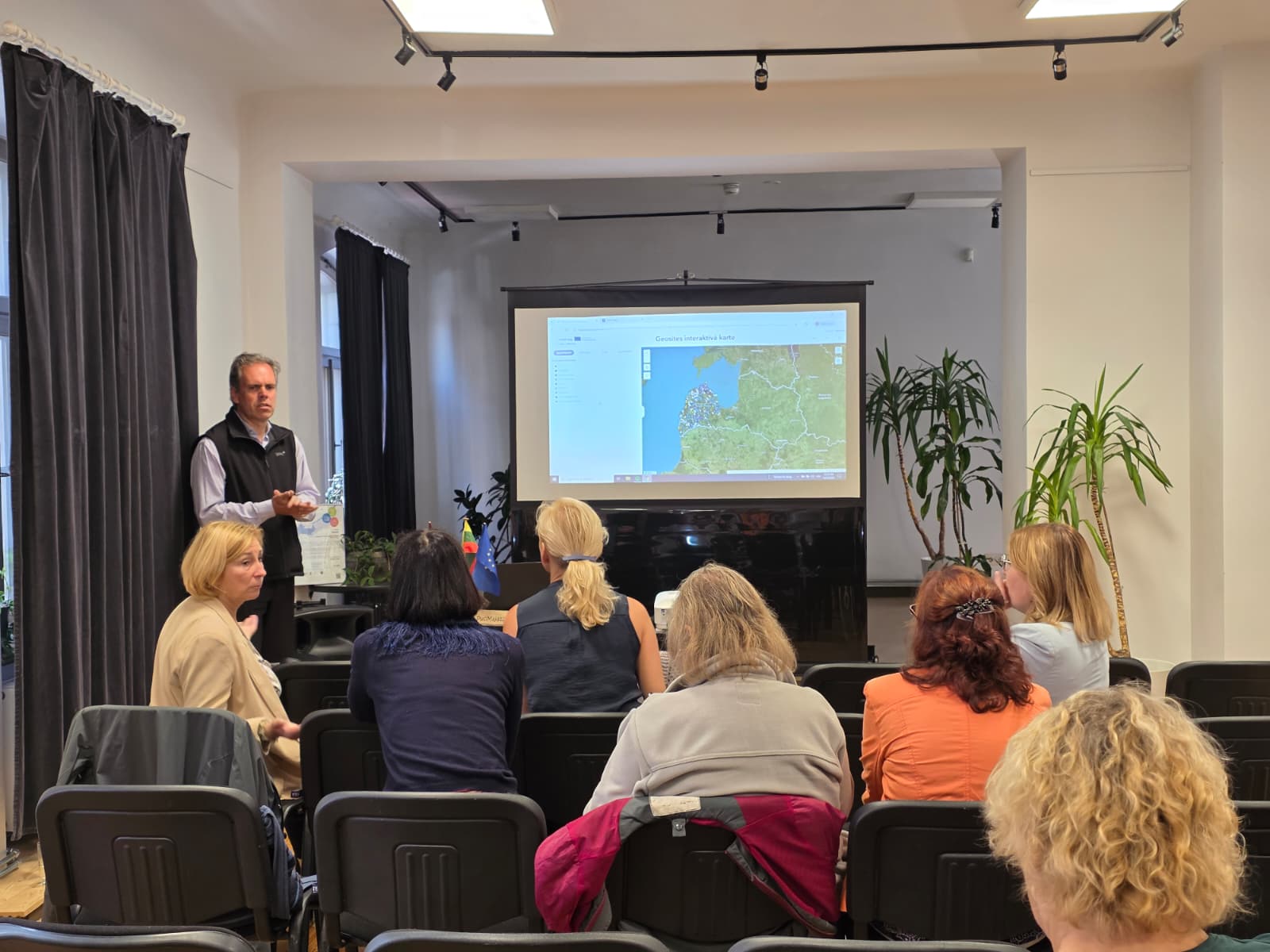 A speaker presents the Geosites database during a seminar in Saldus, with attendees engaged in the discussion. A large screen displays an interactive map, illustrating the practical applications of the database.