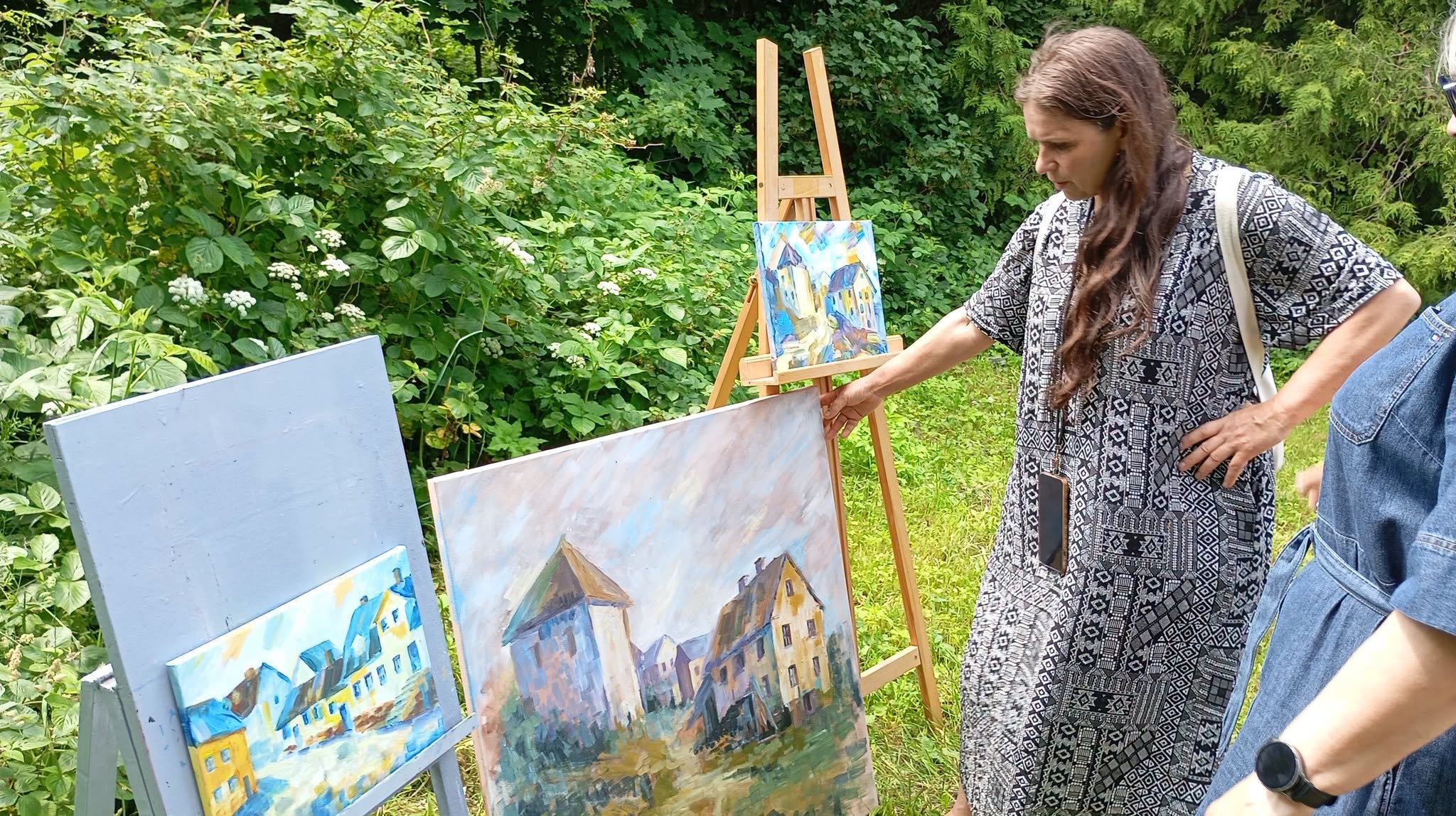 Artist discussing a plein air painting in Kandava, surrounded by vibrant artworks and lush greenery, showcasing collaboration between Latvian and Lithuanian artists.